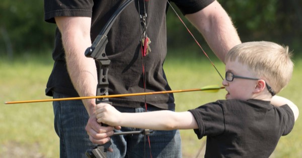 An adult helping a young boy hold a bow correctly while drawing an arrow