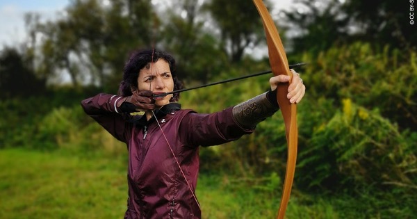 Archer drawing a modern American flatbow (longbow) with a wooden arrow, bow arm extended and draw hand at her face