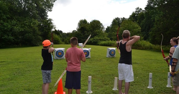 Several people of different ages shooting recurve bows from a grass shooting line towards target bosses at a park archery range
