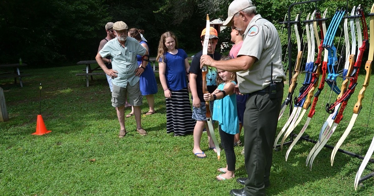 An instructor helping a young archer at a beginner archery session beside a rack of club recurve bows