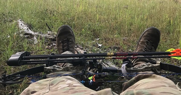 A compound hunting bow with an attached quiver of arrows resting across a bowhunter's lap at the edge of a mountain meadow