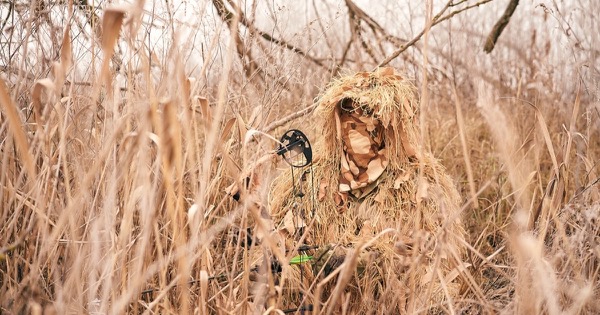 Bowhunter in a grass ghillie suit crouched in tall dry grass with a compound bow