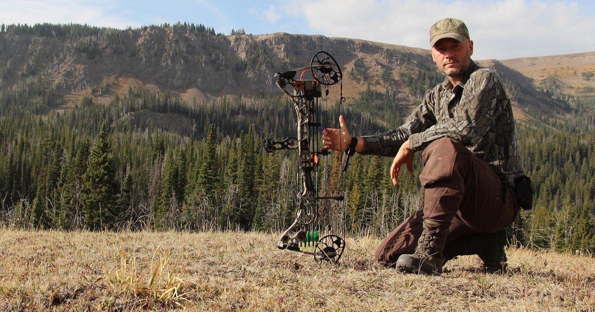 Bowhunter kneeling with a compound bow on a grassy mountain slope above a pine forest