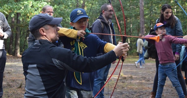 A parent helps a Cub Scout draw a light recurve bow with a soft-tipped arrow at a camporee archery range