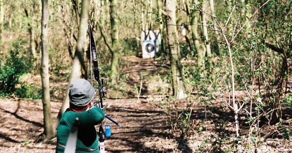 Field archer at full draw with a recurve bow, aiming along a woodland lane at a distant target