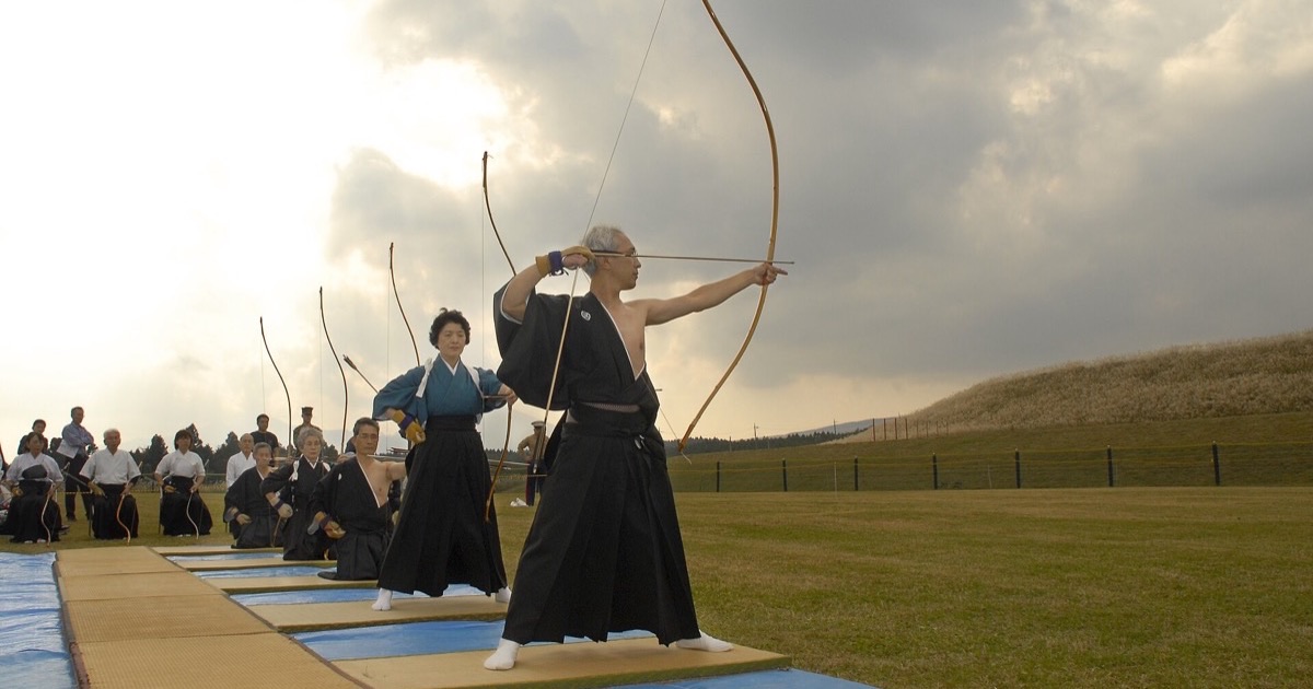 A kyudo archer at full draw with a long asymmetric yumi held below its centre, other archers kneeling behind