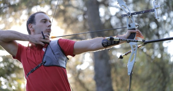 A recurve archer at full draw with a sight and long stabiliser in a wooded setting