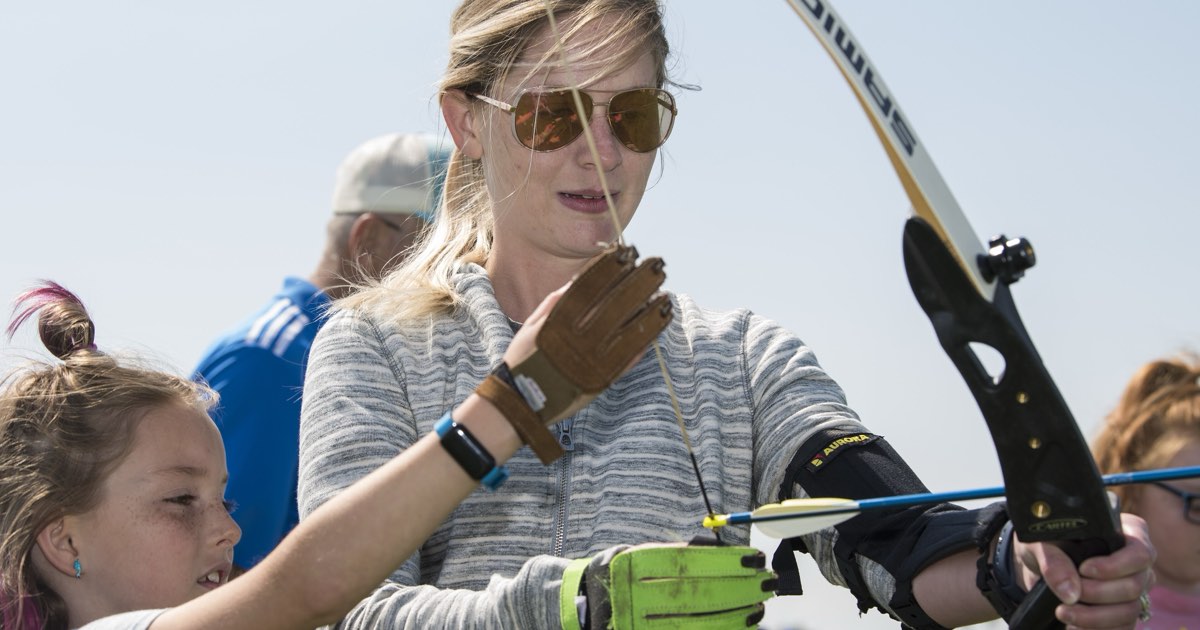 A woman wearing an arm guard and shooting glove holding a recurve bow while helping a child