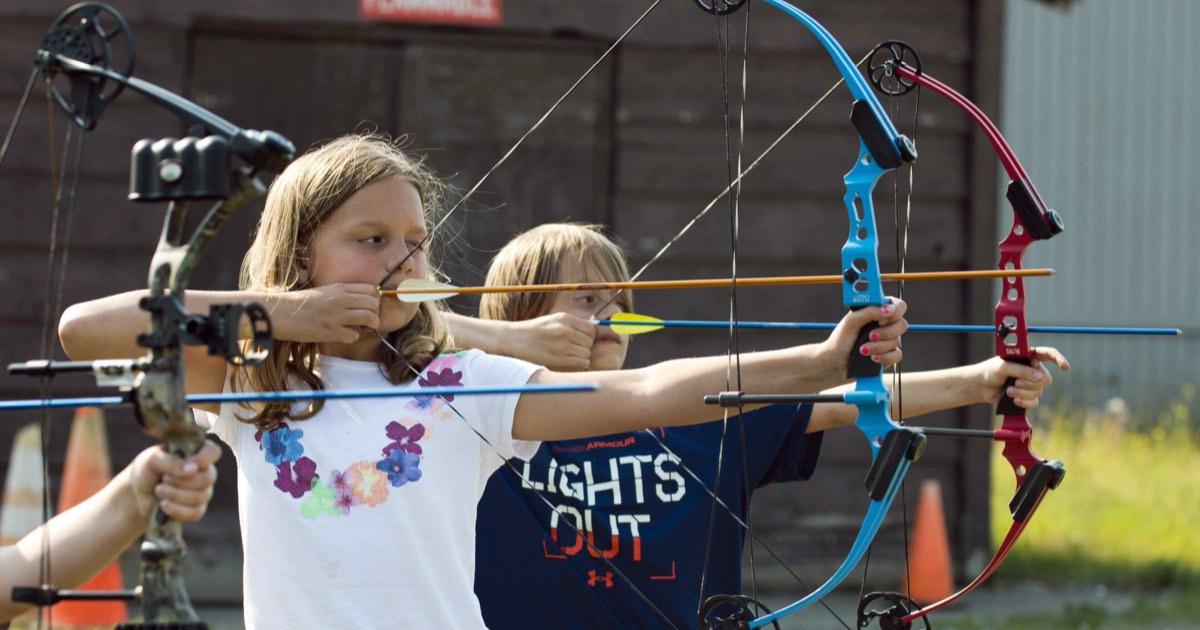 Two young girls aiming youth compound bows side by side at an outdoor range