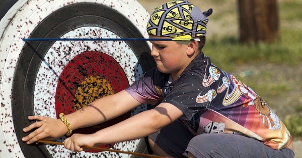 A boy crouches beside a well-used round foam target, holding it with one hand while pulling out an arrow during a youth archery class